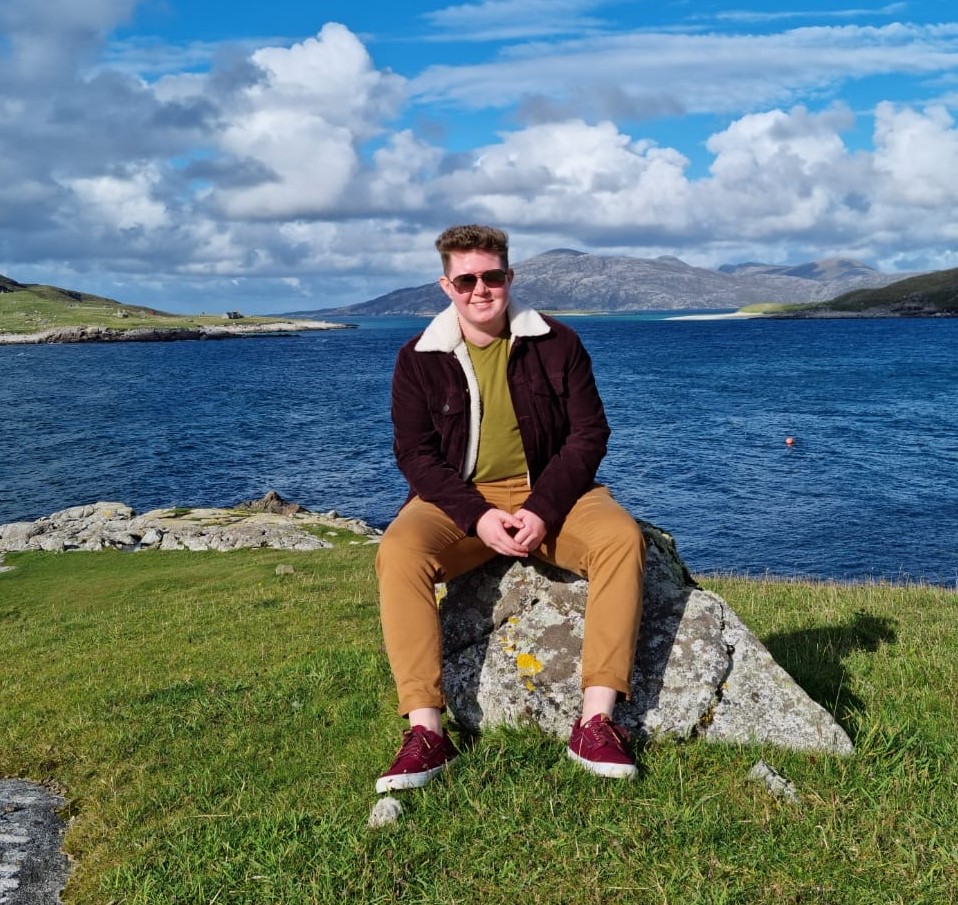 image of Remi dressed in navy shirt, trousers and maroon braces, sat on rainbow steps