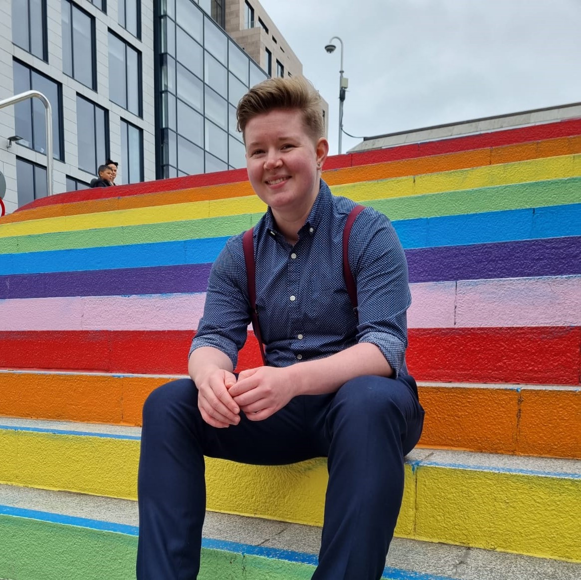 image of Remi dressed in navy shirt, trousers and maroon braces, sat on rainbow steps